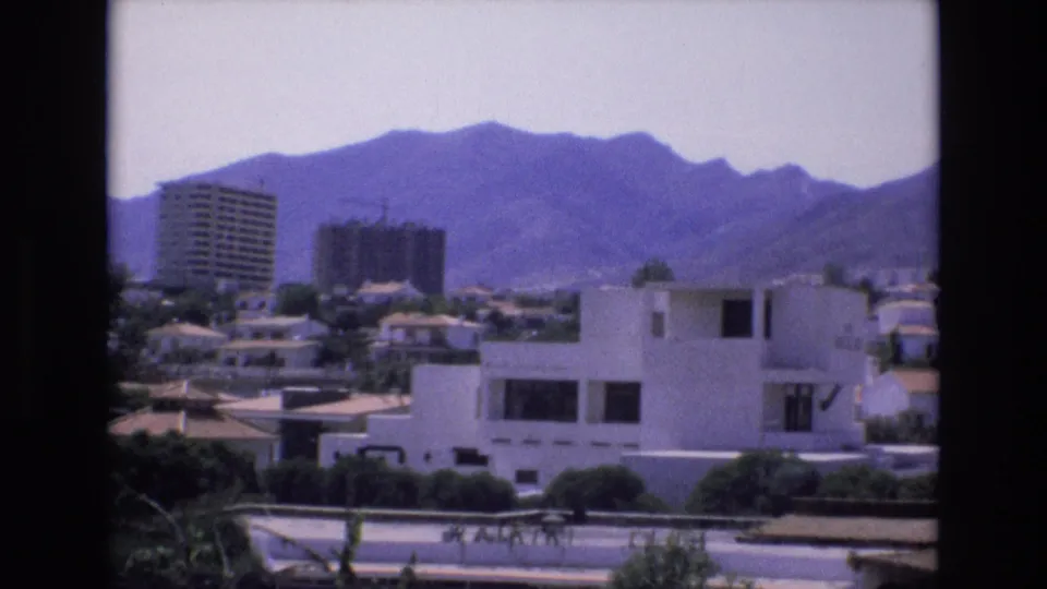 Spain 1972 frame capturing a sunlit residential district with a white flat-roofed building, mid-distance towers, vegetation, and mountains beyond.