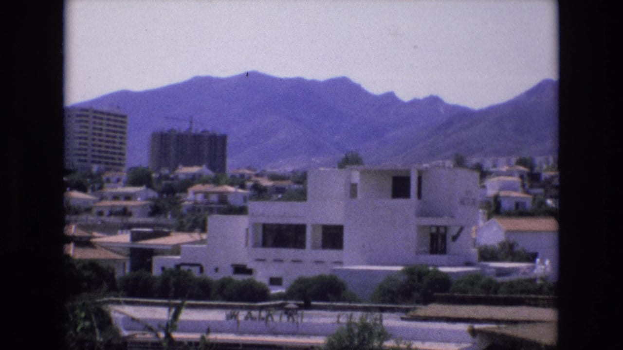 1972 Spain home-movie frame showing white modern residential buildings, tiled roofs, a distant construction tower, and mountains beyond the suburb.