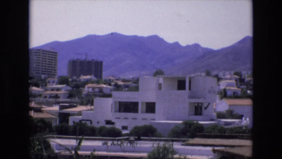 1972 Spain home-movie frame showing white modern residential buildings, tiled roofs, a distant construction tower, and mountains beyond the suburb.