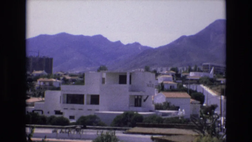 1972 Spain home-movie frame with a white modern house in the foreground, tiled suburban rooftops, mountains, and a construction structure in the distance.