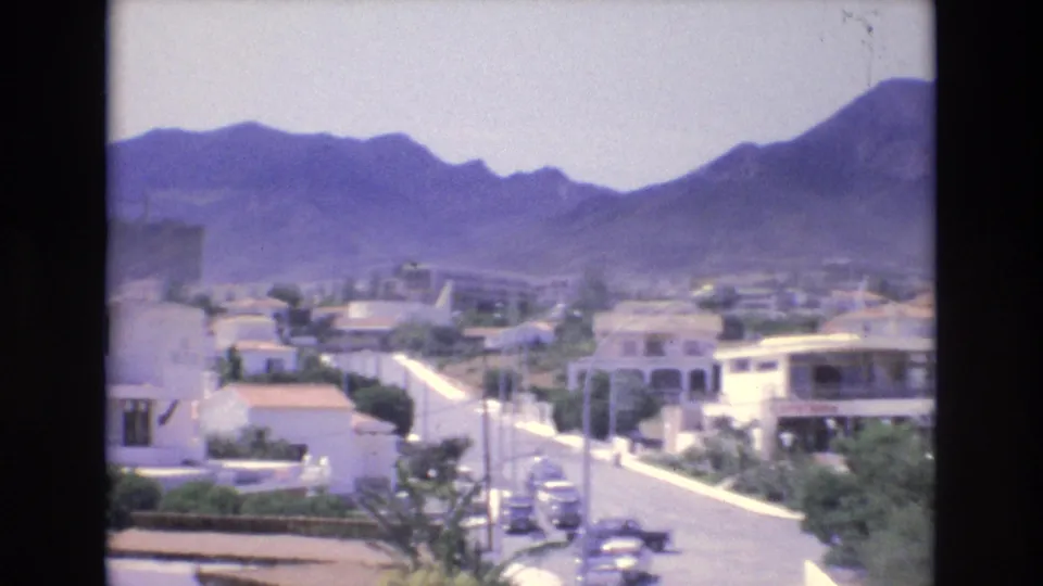 1972 Spain frame showing a bright suburban street lined with white buildings and cars, with mountains rising behind the neighborhood.