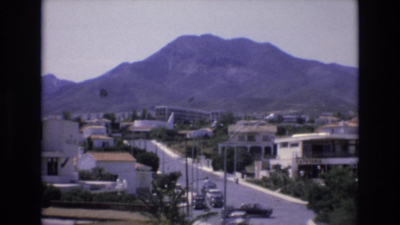 Sunlit residential street beneath a large mountain with white buildings and parked cars.