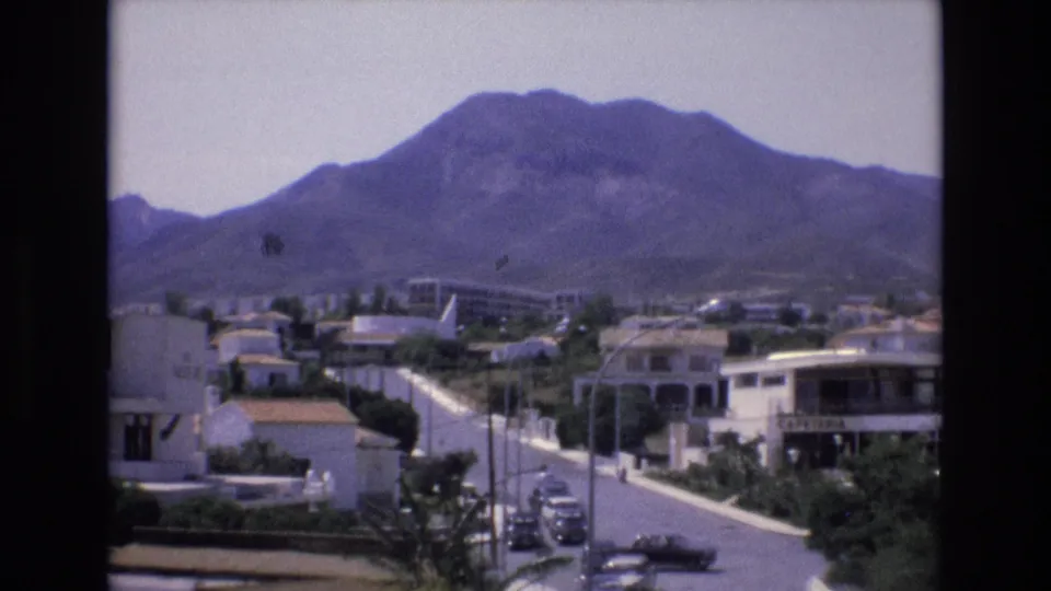 1972 Spain 8mm frame showing a quiet neighborhood street, white-walled buildings, terracotta roofs, parked cars, and a large mountain rising behind the town.