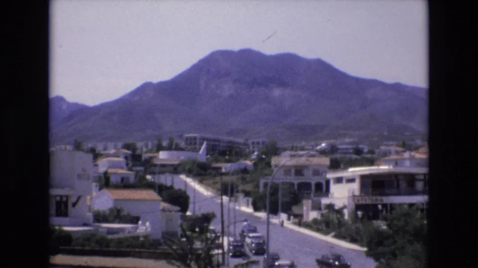 1972 Spain 8mm frame of low-rise homes and a paved street below a broad mountain, with parked cars and bright midday light.