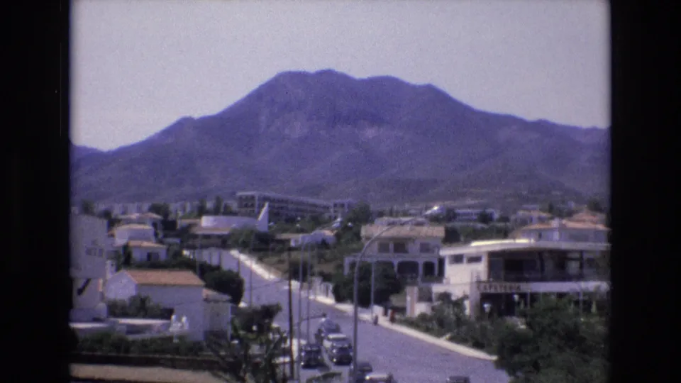 1972 Spain 8mm frame showing white residential buildings, terracotta rooflines, a road with parked cars, and the mountain landscape beyond.