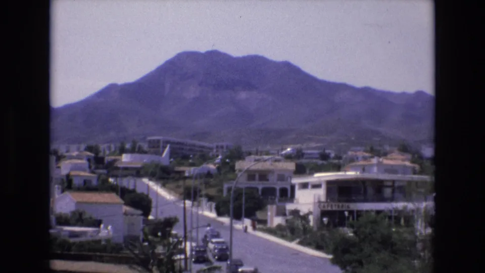 1972 Spain 8mm frame of a quiet Mediterranean neighborhood at the foot of a mountain, with low buildings, parked cars, and roadside greenery.