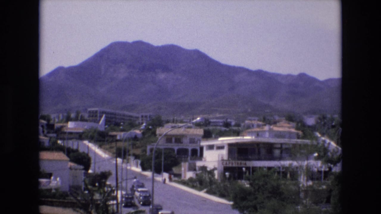 1972 Spain 8mm frame showing a raised view over a curving town road, low white buildings, trees, cars, and a broad mountain ridge.