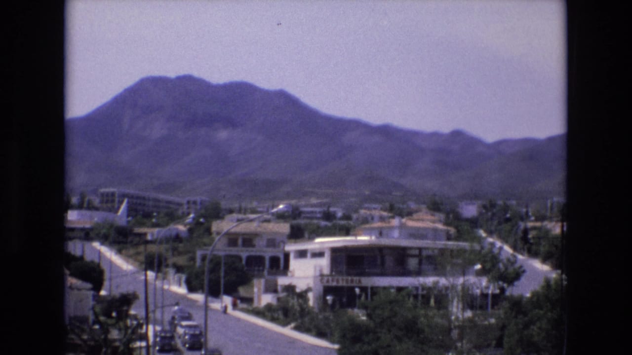 Mountain-backed town view with a road, roadside buildings, trees, and small cars.