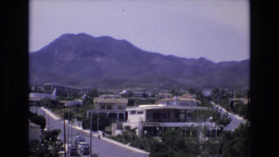 1972 Spain 8mm frame with a steady overlook of low roadside buildings, a curving street, small cars, trees, and mountains in the background.