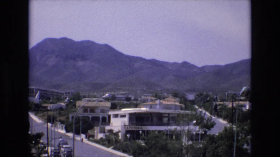 1972 Spain 8mm frame showing a wider elevated view of a sunlit town edge, road traffic, white buildings, vegetation, and a mountain backdrop.