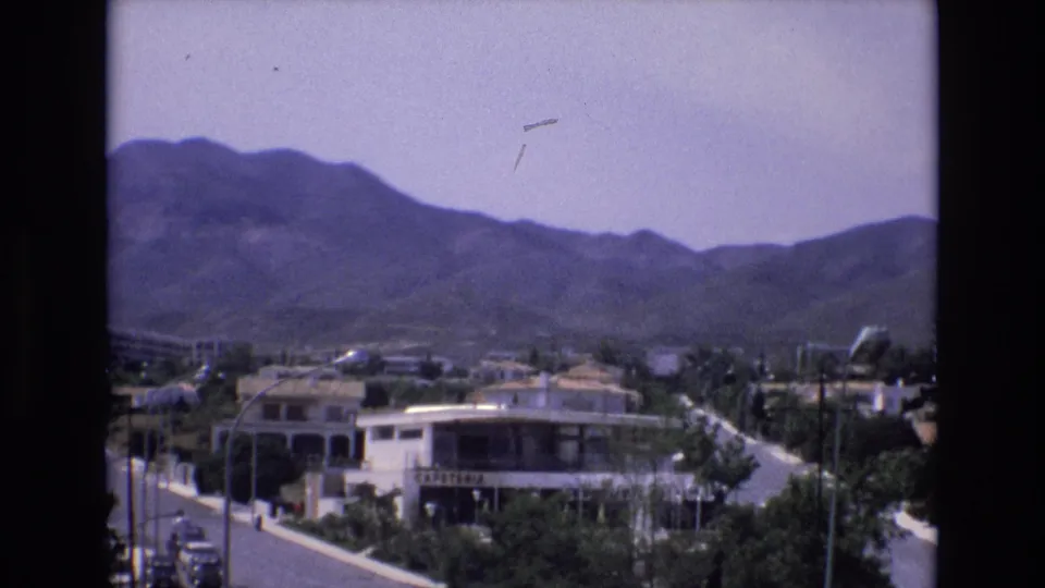 1972 Spain 8mm frame capturing a town street below a dark mountain ridge, with cars, white buildings, trees, poles, and a readable cafeteria sign.