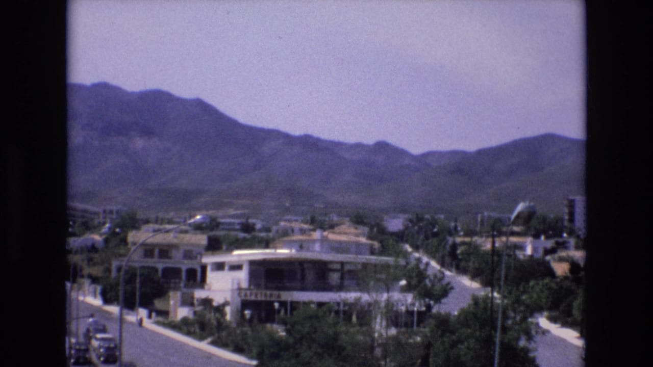 1972 Spain 8mm frame showing an elevated view over a town with a white cafeteria building, paved streets, greenery, scattered cars, and mountains in the background.