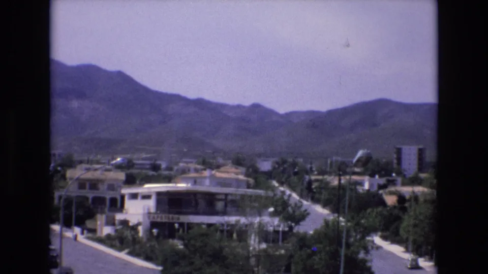 1972 Spain 8mm frame of a mountainside town with a readable cafeteria sign, paved roads, trees, modern low-rise buildings, and distant foothills.