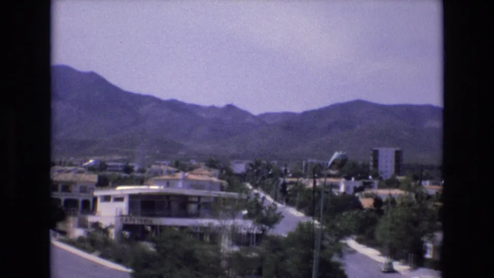 1972 Spain 8mm frame looking across a road and planted town blocks toward a white cafeteria building, residential structures, and mountain foothills.