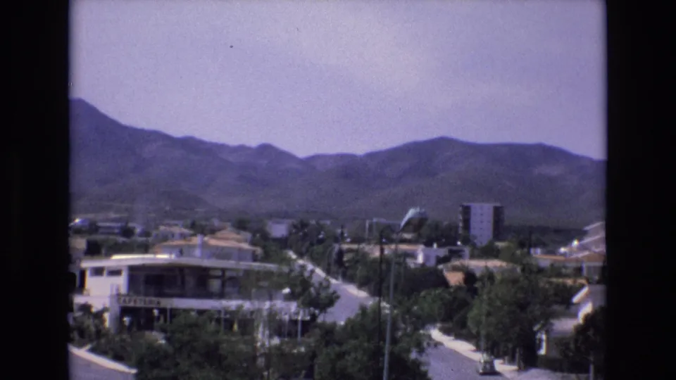 1972 Spain 8mm frame showing a cafeteria-fronted street scene with landscaped greenery, a road with cars, nearby buildings, and foothills beyond the town.