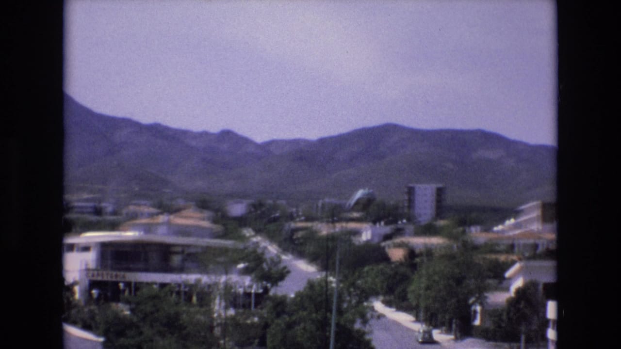 Elevated view of a quiet residential street with buildings, trees, street lamps, and mountains beyond.
