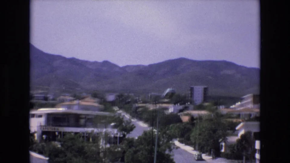 1972 Spain home-movie frame showing a broad residential street below an elevated viewpoint, with low-rise buildings, trees, street lamps, and a mountain range in the distance.