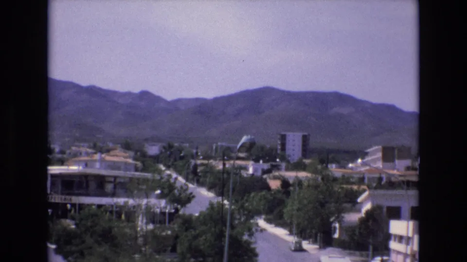 1972 Spain frame with a lightly trafficked residential road, mid-century buildings, trees, a tall apartment block, and a hazy mountain range behind the neighborhood.