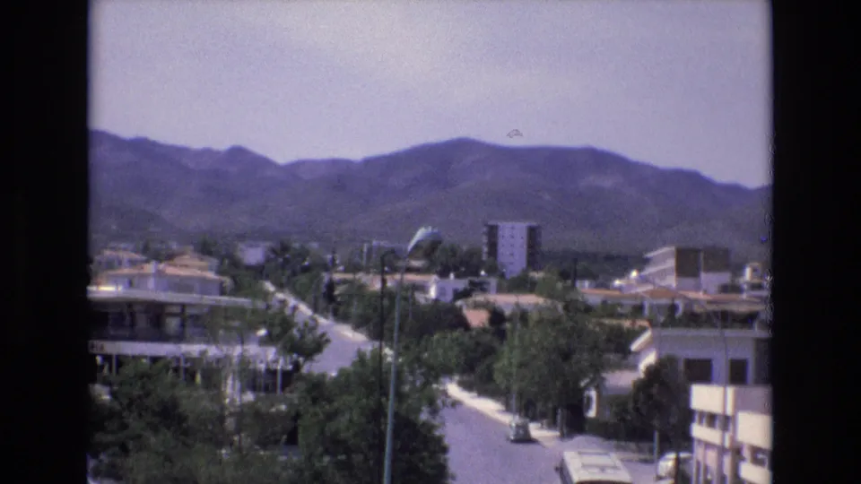 1972 Spain home-movie view of a residential street from above, with a small car on the road, trees and buildings in the foreground, and mountains filling the horizon.
