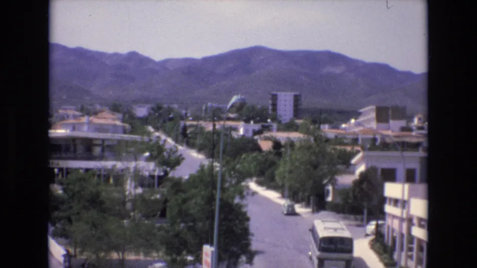 1972 Spain frame showing a quiet residential street beneath a mountain range, with trees, apartment buildings, a small car, and a bus partly visible at the lower right.