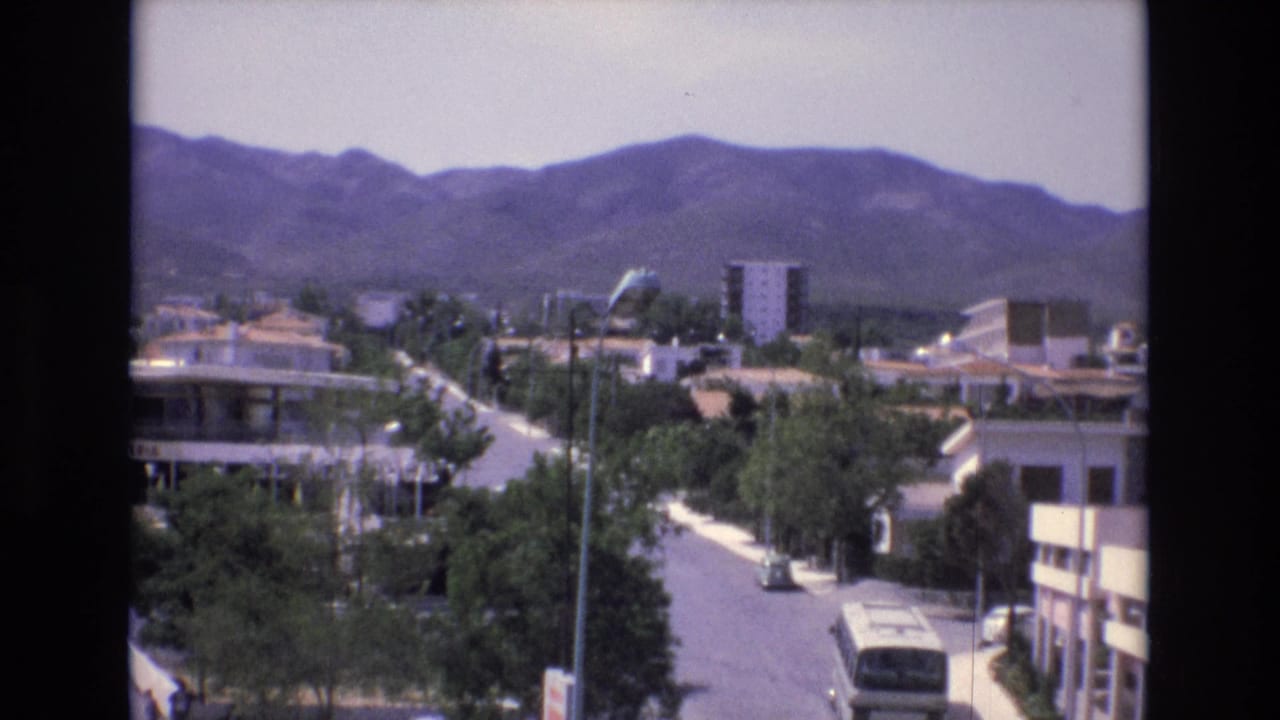 Elevated view of a town street with a bus, low-rise buildings, trees, and hills beyond.