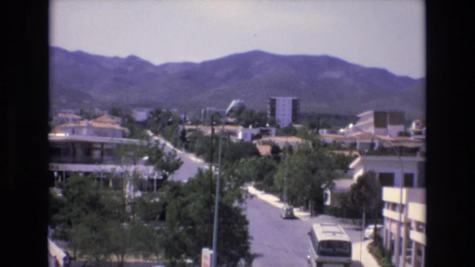 1972 Spain home-movie view from above a quiet street, with a white bus on the road, low-rise buildings, scattered trees, and rolling hills in the distance.