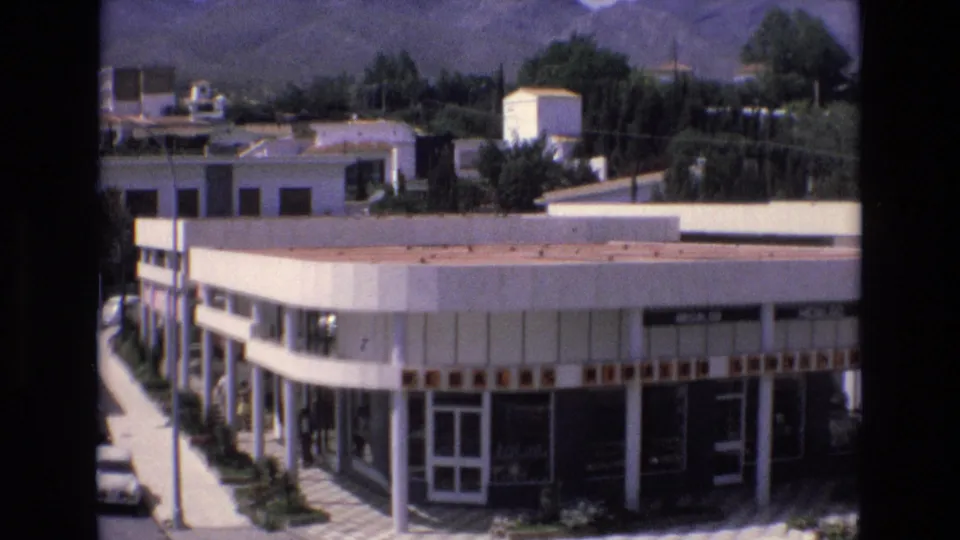 1972 Spain street view showing a flat-roofed commercial building with large windows, a few cars, roadside trees, and hills beyond the town.