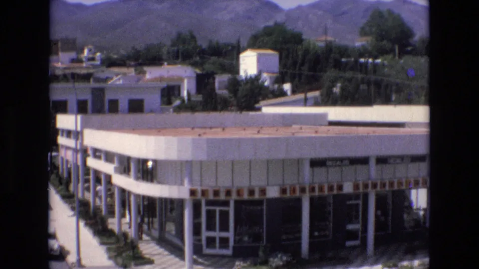 1972 Spain home-movie frame of a corner commercial structure with columns, broad windows, sidewalk shrubs, rooftops, and rolling hills behind it.