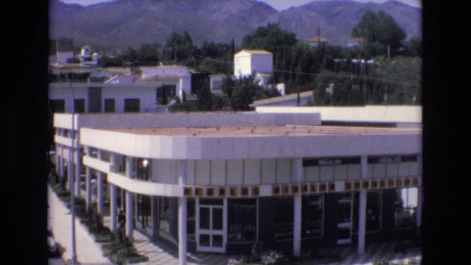 1972 Spain frame showing the same low-rise commercial building from above, with large windows, sidewalk greenery, nearby rooftops, and hills in the background.