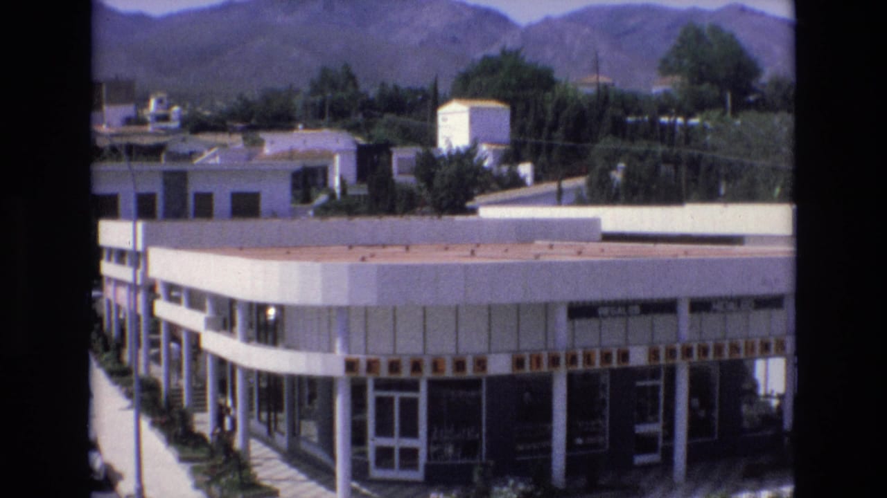 1972 Spain 8mm frame showing a white commercial building with large glass windows, nearby trees, and mountains in the background.