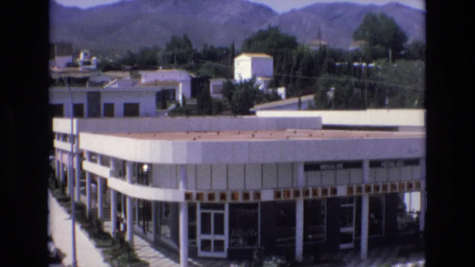 1972 Spain 8mm frame showing a white commercial building with large glass windows, nearby trees, and mountains in the background.