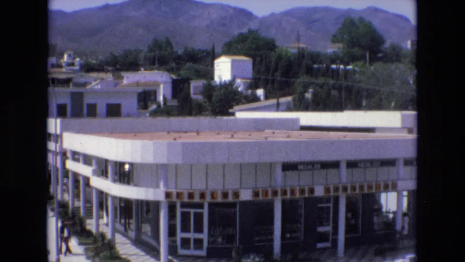 Closer 8mm frame of the white commercial facade in Spain, with glass doors, signage, columns, shrubs, and distant hills visible.