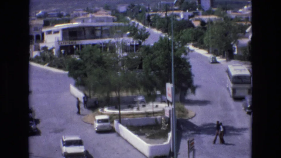 Wider 1972 Spain street view showing a road junction or roundabout with pedestrians, vintage cars, a white bus, trees, and commercial buildings nearby.