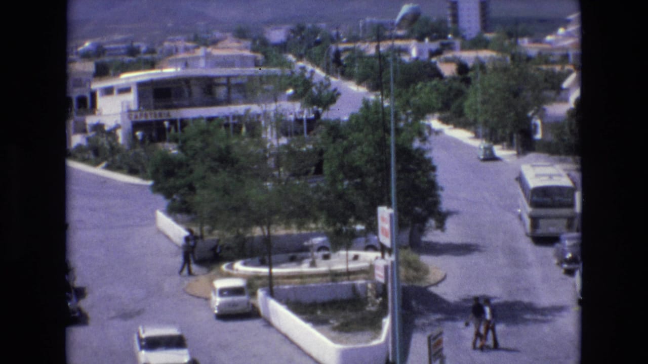 1972 Spain 8mm frame showing an elevated view over a suburban street, with a small round traffic island, low-rise buildings, cars, a bus, trees, and pedestrians crossing in daylight.