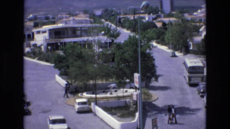 1972 Spain 8mm frame showing an elevated view over a suburban street, with a small round traffic island, low-rise buildings, cars, a bus, trees, and pedestrians crossing in daylight.