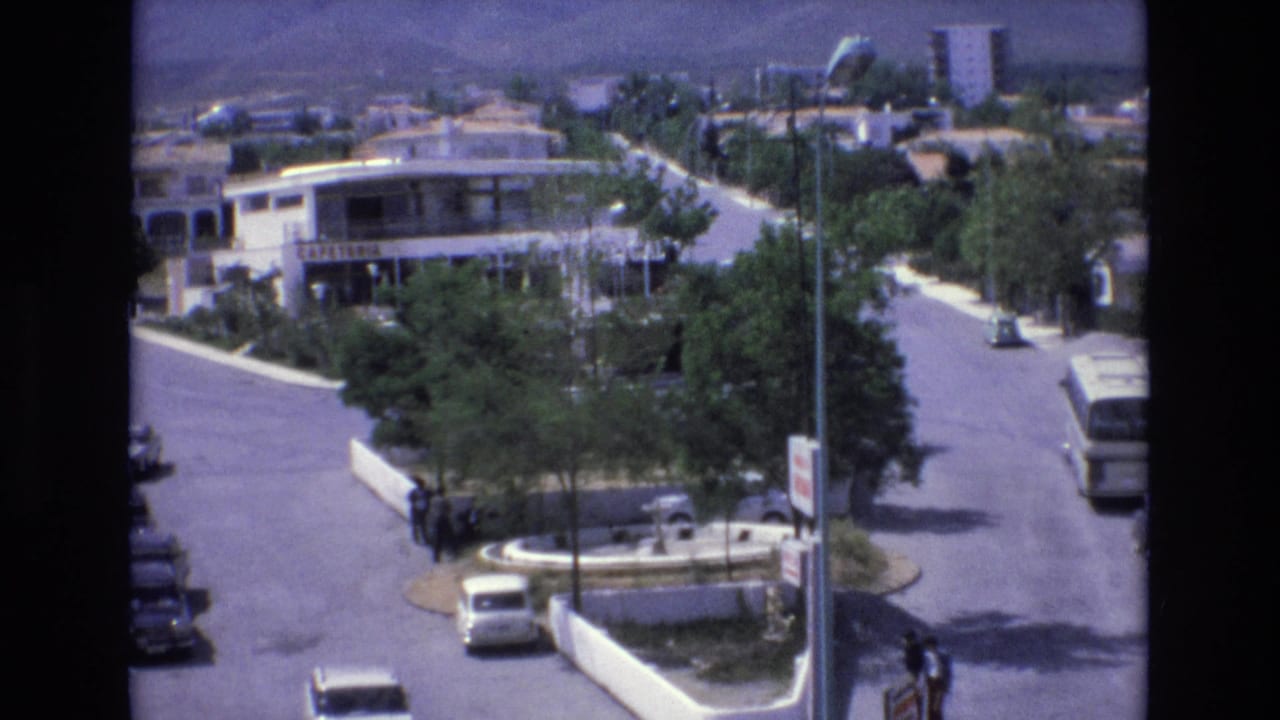 Street corner with a small roundabout, trees, low-rise buildings, pedestrians, cars, and a bus at the road edge.