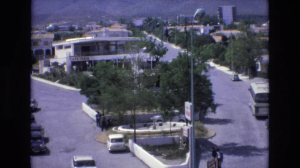 1972 Spain home-movie frame from above a quiet street corner, showing pedestrians near a circular traffic island, several cars, a bus partly at frame edge, trees, and distant hills.
