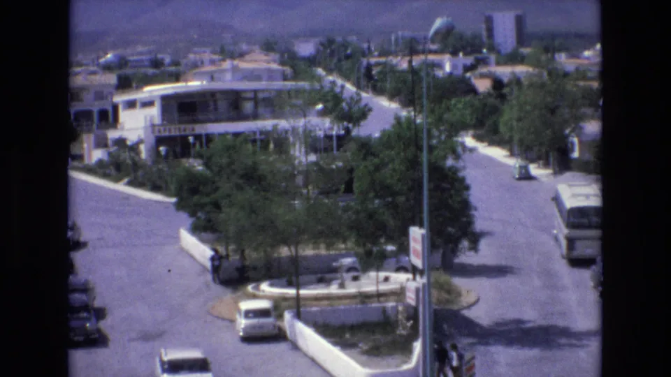 1972 Spain 8mm frame with a low-rise white building, tree-lined road, small traffic island, several 1970s cars, a bus, and pedestrians visible near the street.