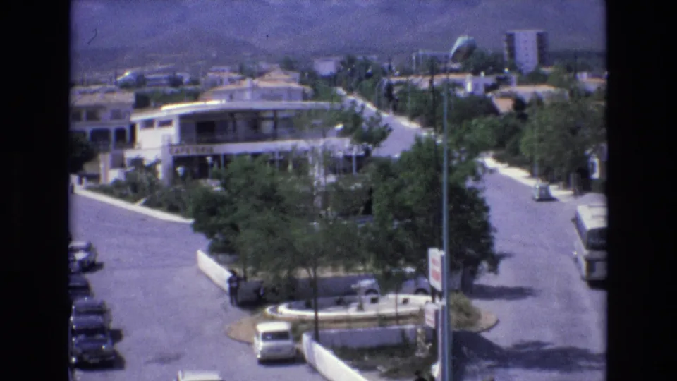1972 Spain home-movie frame showing a suburban road from an elevated angle, with a round traffic island, scattered cars, pedestrians near the crossing, trees, buildings, and hills beyond.