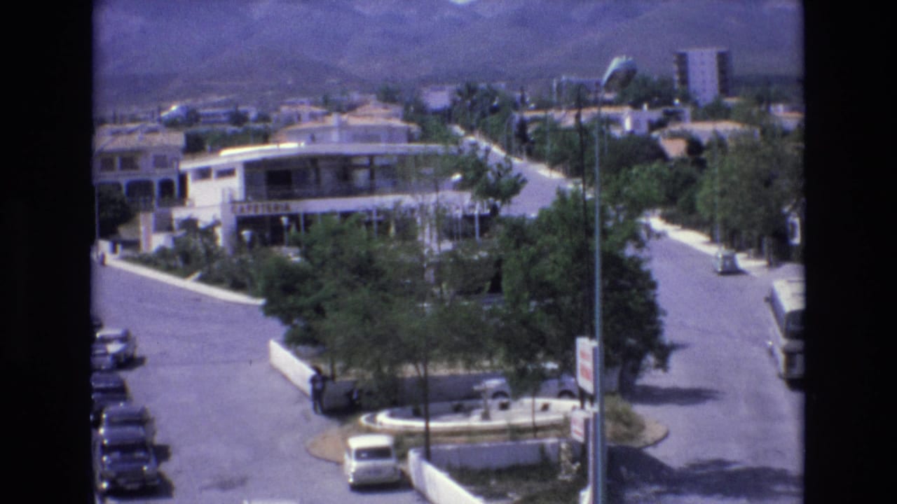 1972 Spain 8mm frame showing an elevated view over a resort street with parked cars, a circular traffic island, trees, a building marked cafeteria, and mountains beyond.