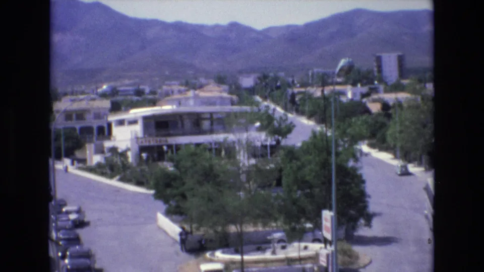 1972 Spain 8mm frame looking across a sunlit resort street with small cars, roadside trees, low buildings, a cafeteria frontage, and a mountainous horizon.