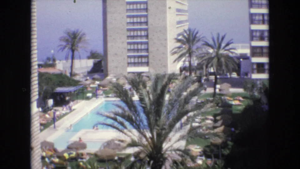 1972 Spain 8mm frame showing an elevated view of a hotel swimming pool with palm trees, umbrellas, lounge chairs, small distant guests, and high-rise hotel buildings.