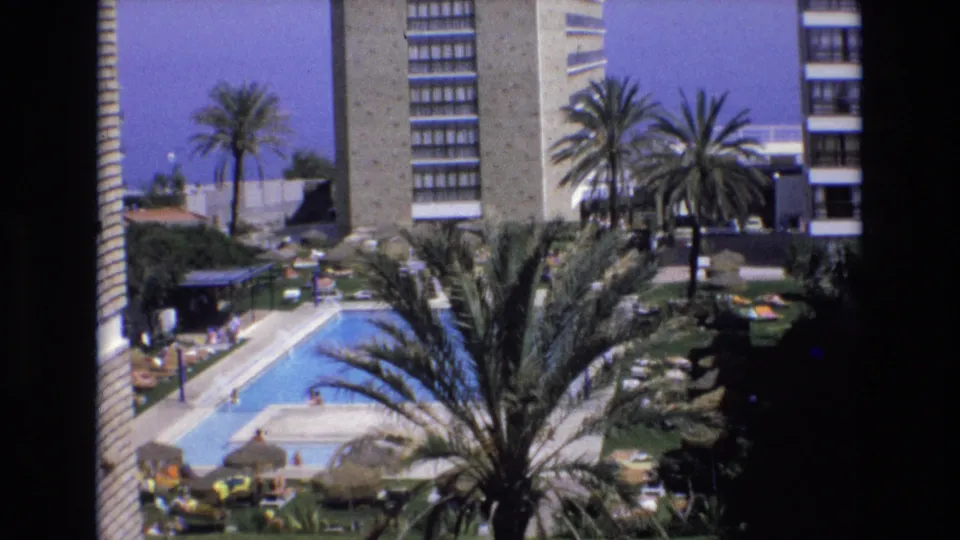 1972 Spain 8mm frame of a resort hotel pool area with a large palm in the foreground, blue swimming pool, umbrellas, distant guests, and multi-story buildings behind.