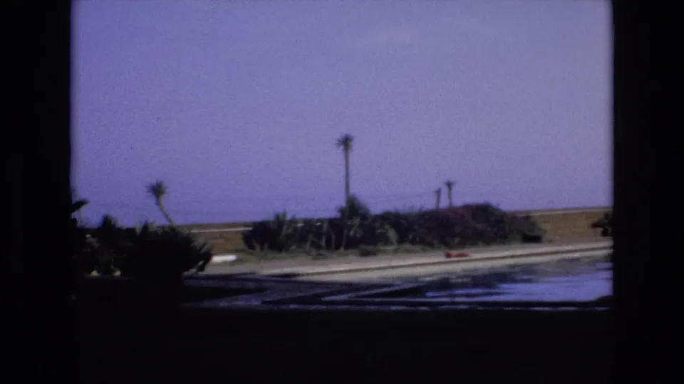 1972 Spain 8mm frame showing a quiet poolside area with reflective water, sparse palms, shrubs, and open ground beyond a low wall.