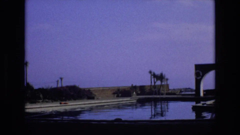 1972 Spain 8mm frame showing the pan reaching a dark arched poolside structure beside reflective water, with palms and open ground behind it.