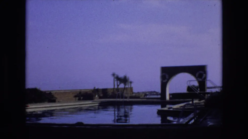 1972 Spain 8mm frame showing a dark arched structure and pool railings at the water's edge, with palms, low wall, and open ground in the background.