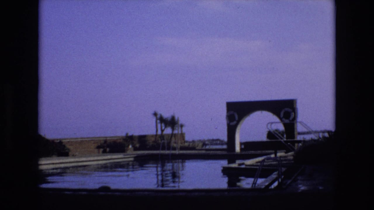 1972 Spain home-movie frame showing a quiet waterfront with reflective water, a small dock, palm trees, and a distinctive arched structure in the distance.