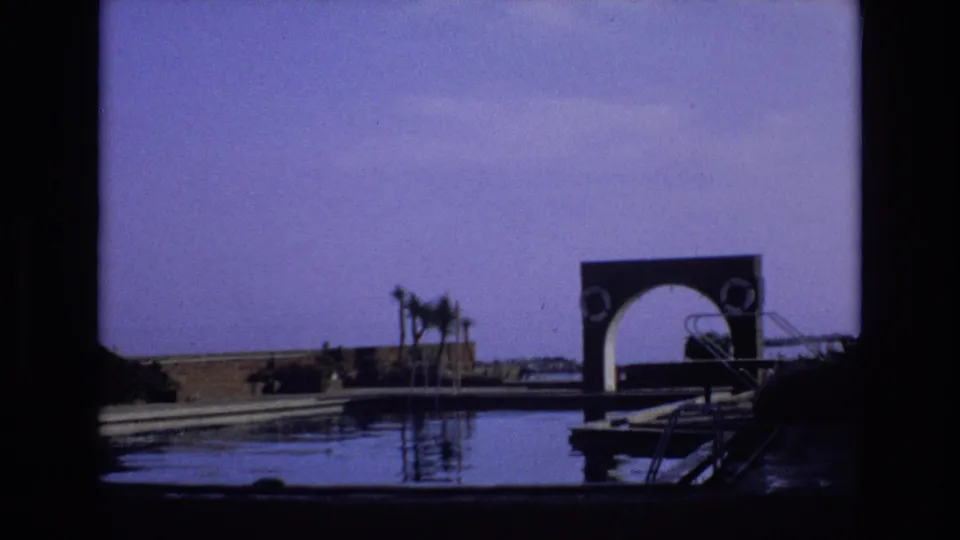 1972 Spain home-movie frame showing a quiet waterfront with reflective water, a small dock, palm trees, and a distinctive arched structure in the distance.