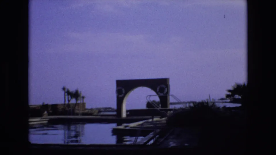 1972 Spain home-movie frame showing reflective water, a low dock, palms, and a large arched structure seen through dark foreground borders.