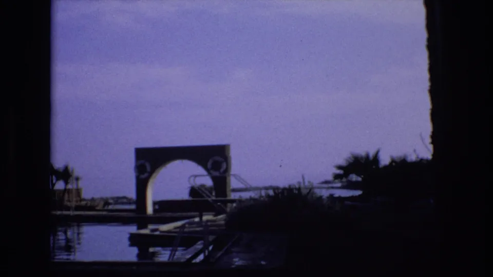 1972 Spain frame with a partially shadowed foreground framing calm water, dock elements, palms, and the same arched waterfront structure.